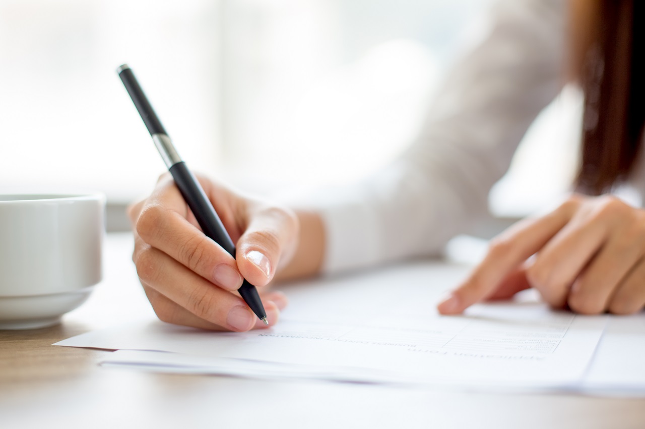 Hand of young businesswoman writing on paper or signing contract at table in office